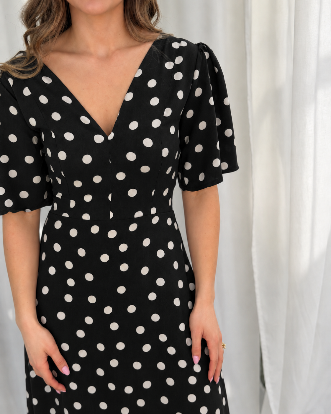 A woman wearing a black dress with white polka dots, featuring a V-neckline, short puff sleeves, and a fitted waist, posed against a white curtain background.