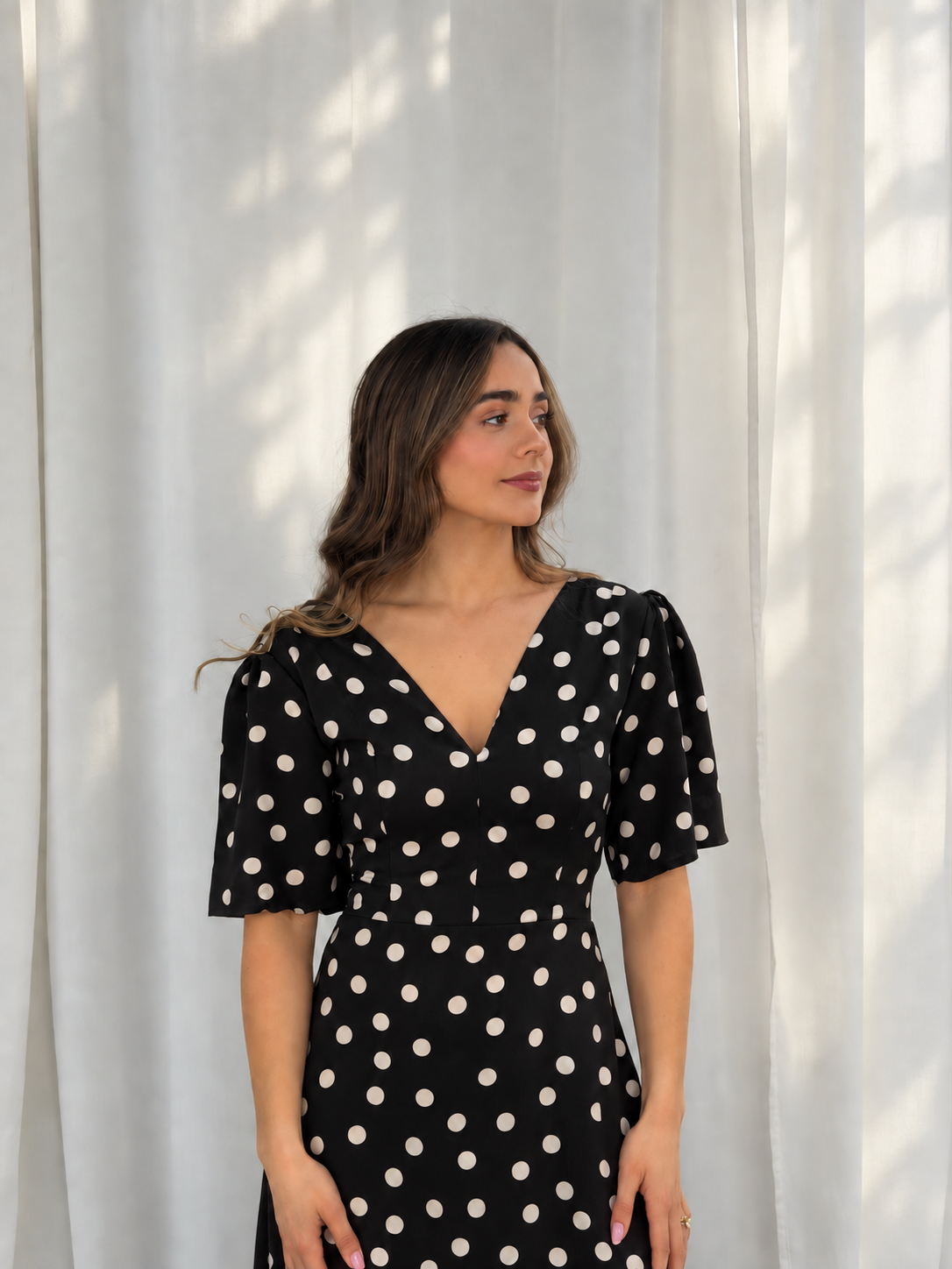 A woman wearing a black V-neck dress with white polka dots, standing in front of a white curtain backdrop.