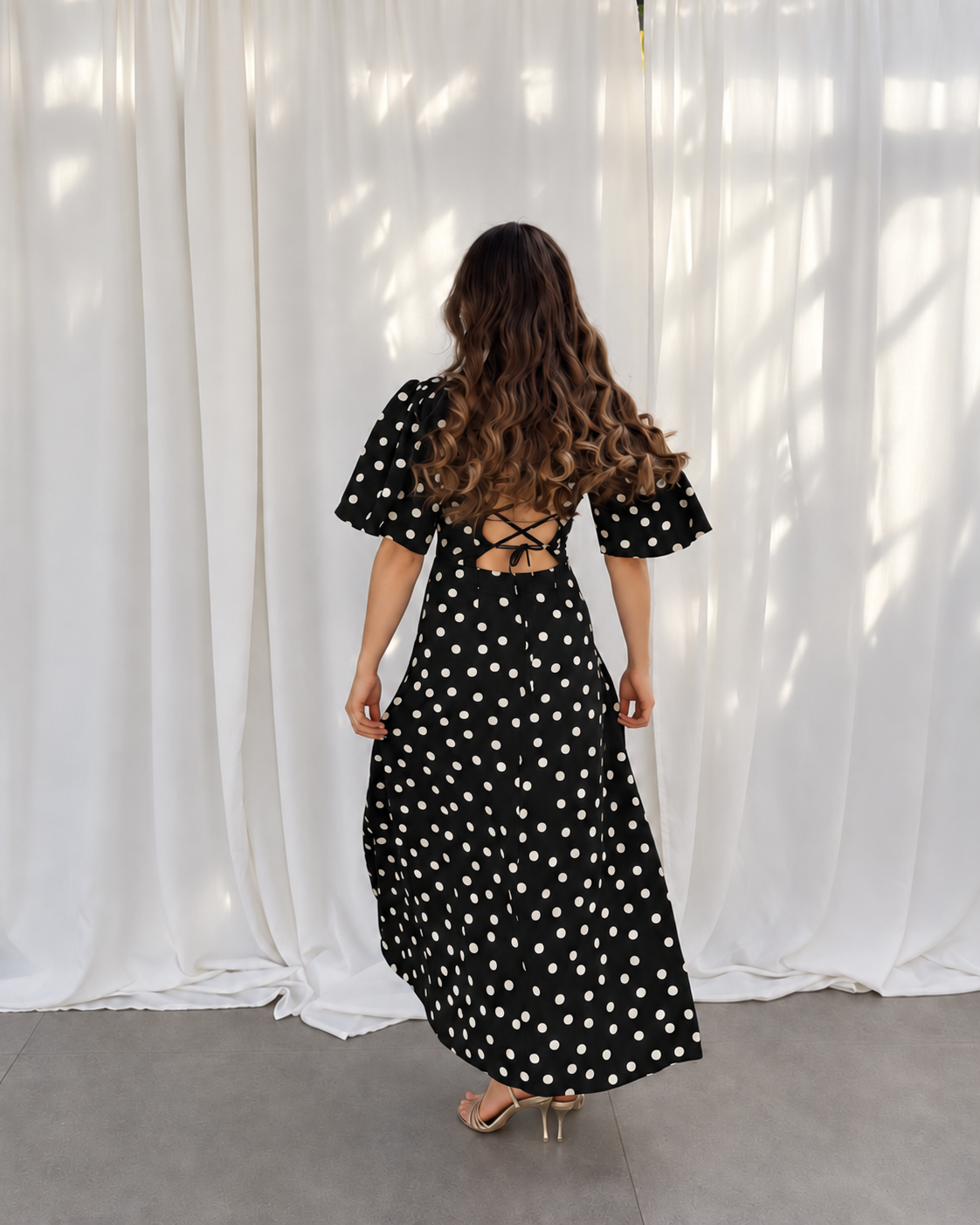 A woman viewed from behind wearing a black midi dress with white polka dots, puff sleeves, and a lace-up open back, standing in front of a draped white curtain.